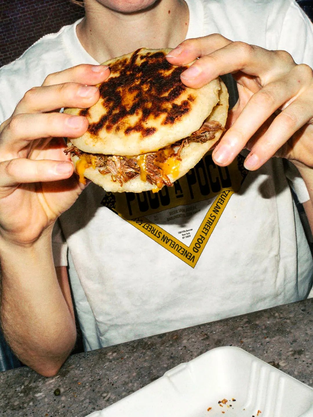 person wearing Poco A Poco T-shirt eating an Arepa